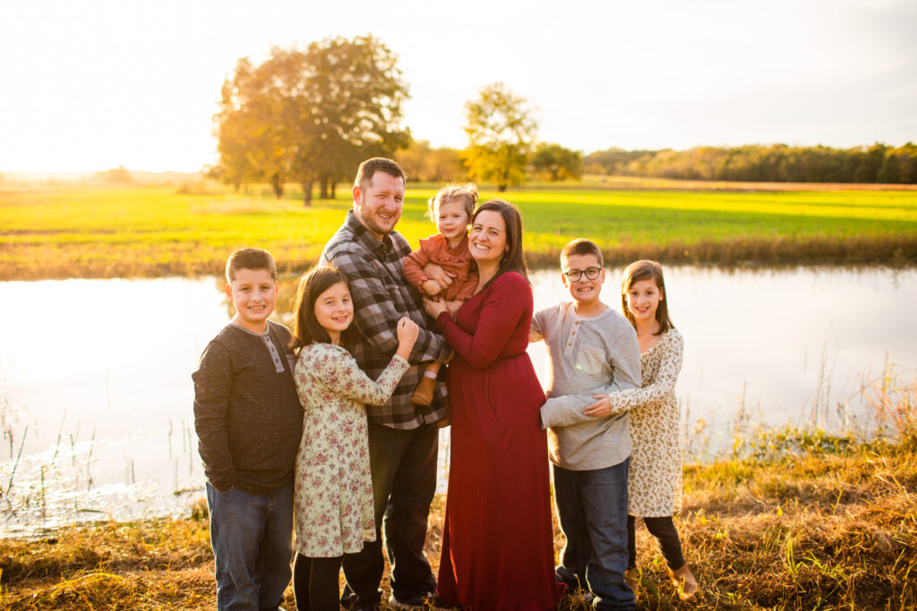 Photo of a happy family near a lake. all of the kids are smiling. The parents are holding one of the kids.